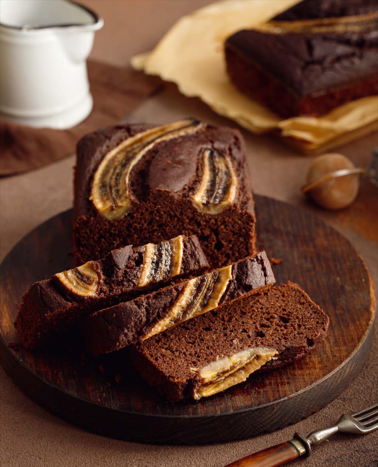 Chocolate banana bread sliced on a wooden board with a fork and a white container in the background.