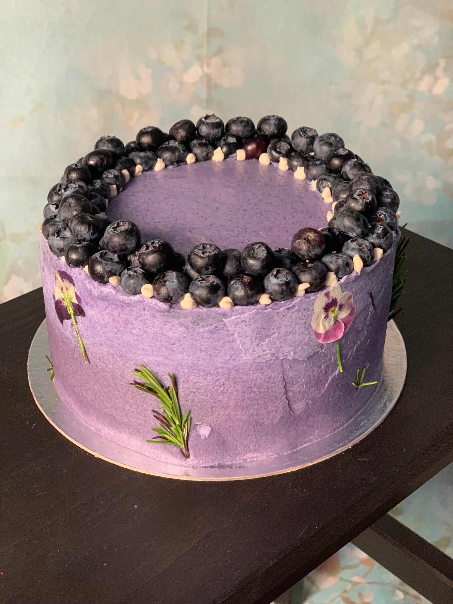 A purple vegan blueberry matcha cake with a layer of blueberries on top, set on a wooden stand with a floral pattern in the background.
