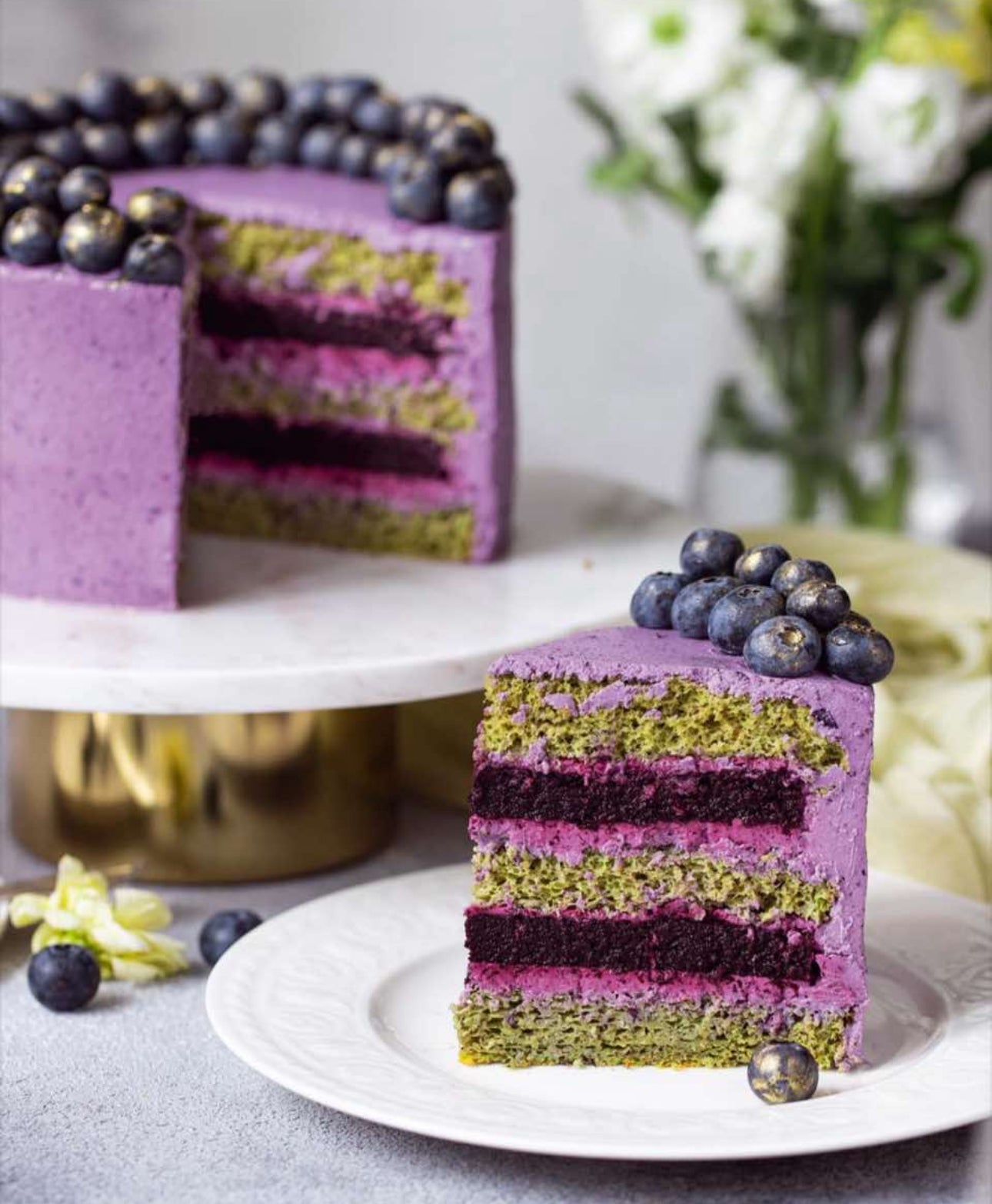 Layered cake with purple, green, and pink layers on a white plate, with a full cake in the background.