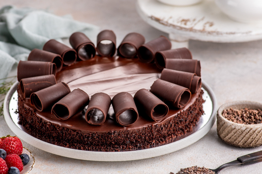 Chocolate cake with chocolate curls on a white plate, surrounded by berries and a bowl of chocolate shavings.