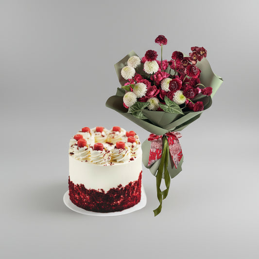 Red velvet cake with white frosting and red berries next to a bouquet of red and white flowers on a light gray background