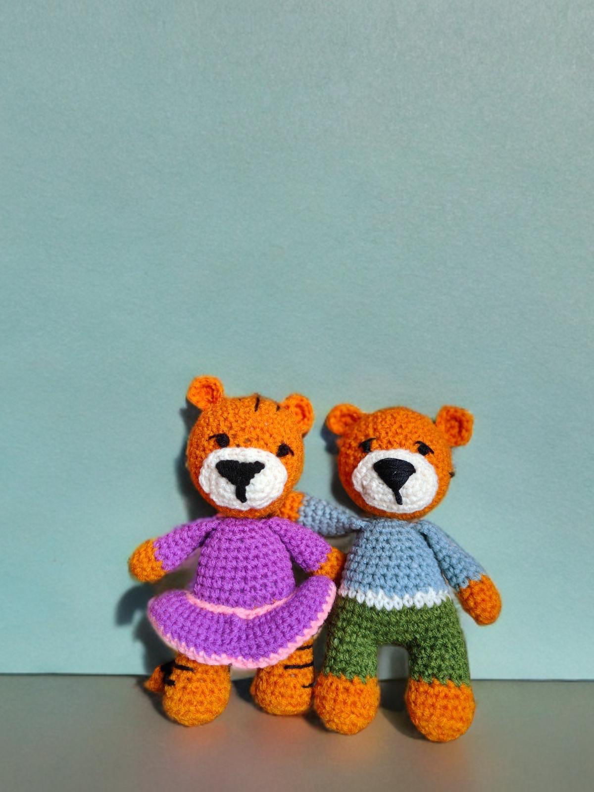 Two crochet teddy bears gifts, one in a dress and one in pants, against a light blue background.