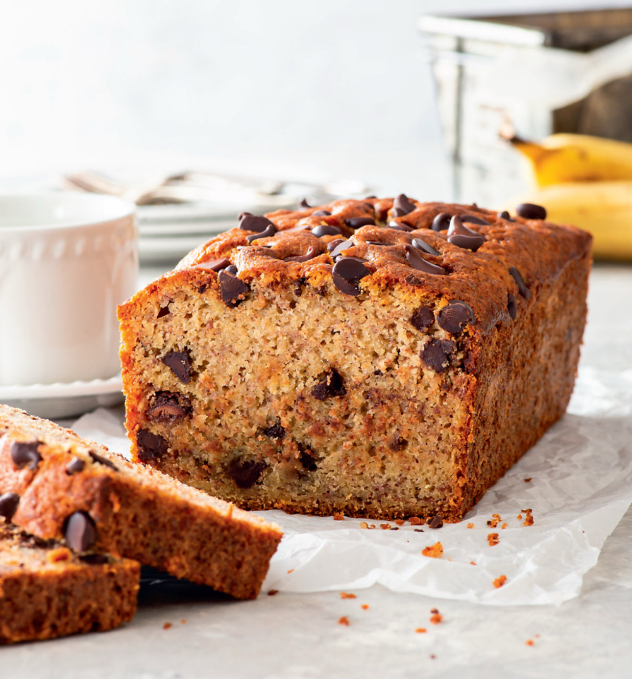 Loaf of banana bread with chocolate chips on a white surface, with a cup and bananas in the background.