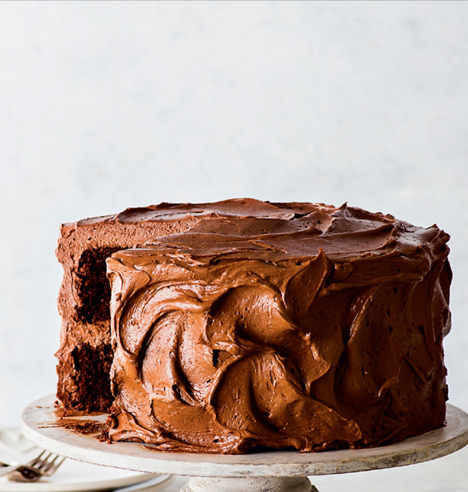 A gluten-free chocolate cake with a frosting layer and chocolate sprinkles on top, placed on a white cake stand against a light background.