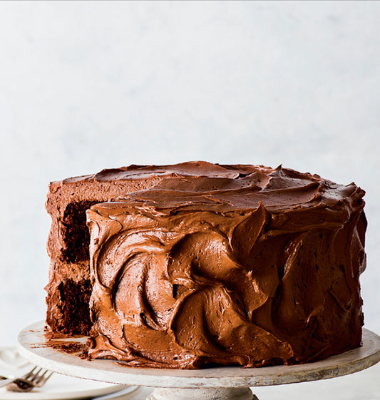 A gluten-free chocolate cake with a frosting layer and chocolate sprinkles on top, placed on a white cake stand against a light background.