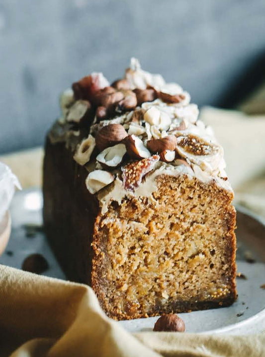 A gluten-free vegan carrot loaf with a layer of chopped nuts and dried figs on top, placed on a plate with a piece of almond flour bread in the background.