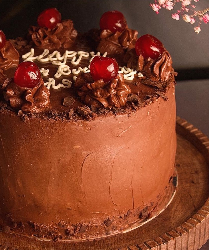 A decorated chocolate sponge cake with cherries and the words 'Happy Birthday' written on top in a white cream, garnished with glazed cherries on the side.
